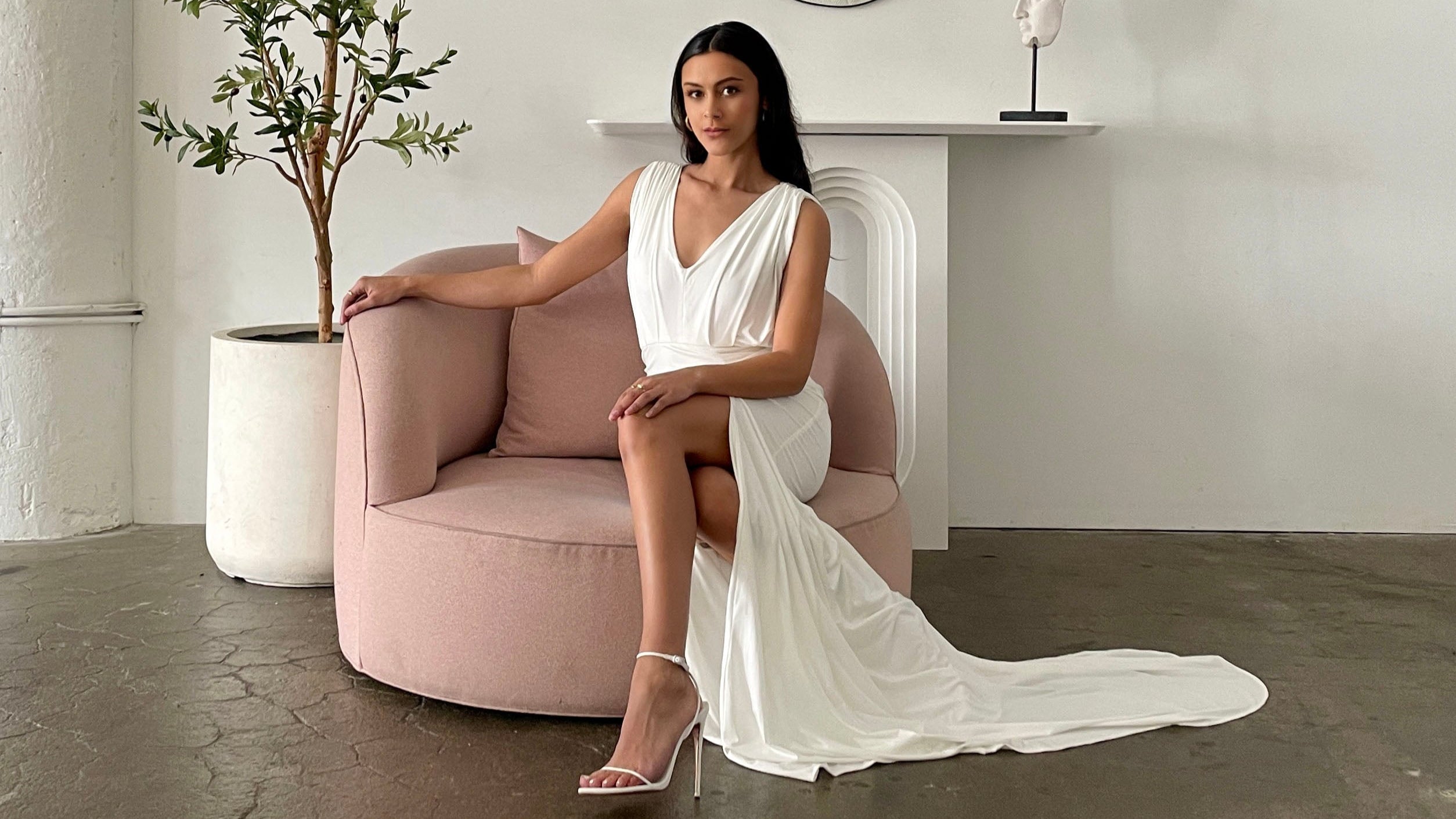 Woman in a white simple wedding dress sitting on a pink chair in a minimalistic room.